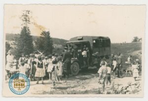 Grupa dzieci w strojach ludowych z żołnierzami przed samochodem wojskowym