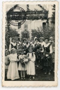 Grupa kobiet w strojach ludowych i dzieci przed bramą witającą biskupa