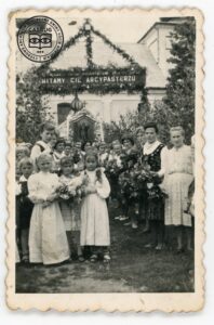 Grupa kobiet w strojach ludowych i dzieci przed bramą witającą biskupa